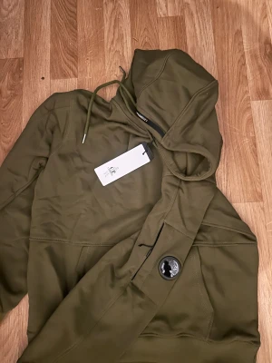 Olivgrön hoodie från C.P. Company - Snygg olivgrön hoodie från C.P. Company med huva och snörning. Tröjan har en cool detalj med en svart lins på ärmen och ribbade muddar. Perfekt för dig som gillar streetwear och vill ha något unikt i garderoben.