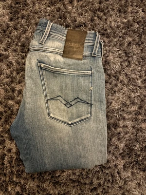 Replay Anbass ljusblå  - Snygga Replay Anbass jeans i ljusblå tvätt med slitningar och slim passform. Klassisk femficksdesign, coolt broderi på bakfickan och detaljerade slitningar framtill. Perfekt för dig som gillar en modern och avslappnad stil.