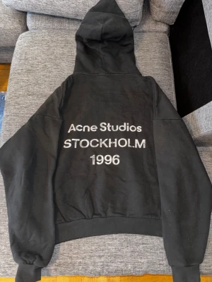 Svart hoodie Acne Studios S - Svart hoodie från Acne Studios i storlek S med tryck på ryggen: 'Acne Studios STOCKHOLM 1996'. Tröjan har huva, långa ärmar och är tillverkad i 100% bomull med mjuk insida. Perfekt för dig som gillar stilren streetwear med svensk design.