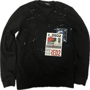   Dsquared2 Tröja - Svart sweatshirt från Dsquared2 med coola tryck och patchar framtill, samt handskrivna detaljer och färgstänk i blått och vitt över hela tröjan. Rund hals och långa ärmar, tillverkad i mjuk bomull. Storlek är M men passar S.