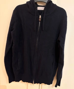 Mörkblå hoodie i kashmir från Sayless - Mjuk och lyxig mörkblå hoodie från Sayless i 100% kashmir. Tröjan har huva, långa ärmar och praktisk dragkedja framtill. Perfekt för dig som gillar stilrena och bekväma plagg med exklusiv känsla. Snygga fickor på sidorna och ribbad kant nedtill.