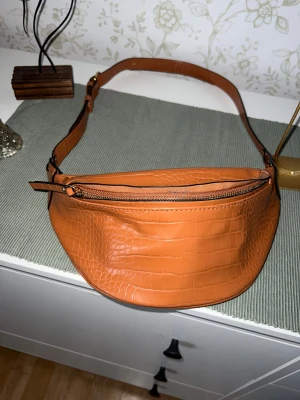 Brun axelväska i croco-imitation - Snygg brun axelväska i croco-imitation med justerbart axelband och dragkedja upptill. Väskan har ett rymligt huvudfack och en innerficka med dragkedja. Perfekt för att bära dina viktigaste grejer med stil.