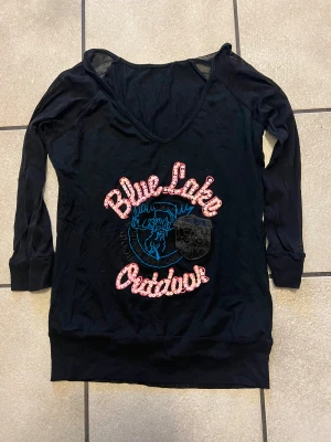 Svart långärmad tröja Blue Lake storlek XS/S - Svart långärmad tröja från Blue Lake med mesh-ärmar och v-ringad hals. Framsidan har ett stort tryck med blå älg och text i rosa paljetter. Patch med siffran 38 på bröstet. Tröjan är loose fit och har bred mudd nedtill.