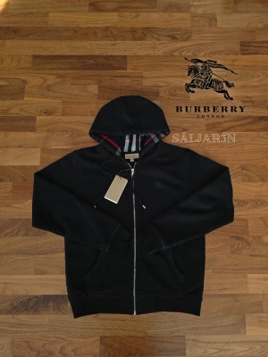 Svart Burberry Hoodie - ❗️Black Friday Erbjudande❗️                                Letar du efter en stilren och lyxig hoodie? Denna helt nya grå Burberry-hoodie kombinerar elegans med hög komfort. Den har Burberrys ikoniska rutmönster i fodret och en diskret logga på bröstet för en exklusiv touch.  Metallbiten är trådanra är borta. Tillverkad av mjukt, slitstarkt material och utrustad med dragkedja samt huva – både praktisk och stilfull. Passar som M/L.📌 Skick: Ny med taggar. 📦 Snabb leverans – skickas samma dag!