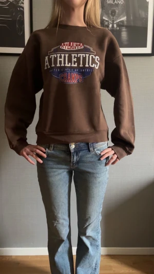 Brun tjocktröja med tryck🤎 - Brun tjocktröja med stort Athletics-tryck i vitt, blått och rött. Tröjan har rund hals, långa ärmar och ribbade muddar. 