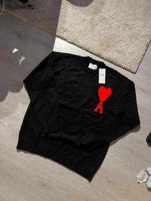 Svart stickad tröja från Ami Paris - Svart stickad tröja från Ami Paris med ett stort rött hjärta och bokstaven A broderat på bröstet. Tröjan har rund hals, ribbade muddar och lång ärm. Materialet är mjuk ull som känns skön mot huden. Perfekt för dig som gillar stilrena plagg med en twist.