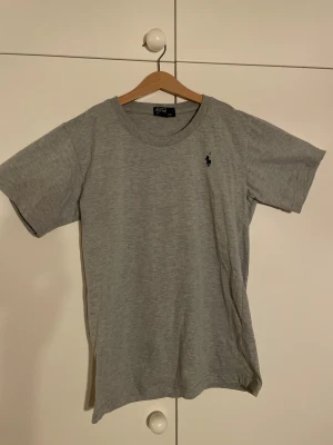 Grå t-shirt Polo Ralph Lauren - Snygg grå t-shirt från Polo Ralph Lauren med klassisk rund hals och korta ärmar. T-shirten har en liten svart Polo-logga broderad på bröstet. Perfekt basic med stilren look.