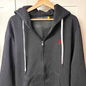 Svart hoodie från Polo Ralph Lauren - Svart hoodie från Polo Ralph Lauren med röd broderad logga på bröstet. Tröjan har vit snörning i huvan, dragkedja framtill och två fickor. Tillverkad i mjuk bomull, perfekt för chill dagar. Klassisk och stilren design som alltid funkar. Storlek M men skulle kunna passa för både S/xs