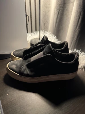 Svarta sneakers från Axel Arigato - Svarta sneakers från Axel Arigato med minimalistisk design och vit platt sula. Skorna har elastisk panel istället för snörning och är tillverkade i textil och skinn. Perfekta för dig som gillar stilrena och enkla skor med en modern touch.