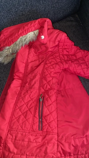 Röd quiltad pufferjacka med fuskpäls - Snygg röd pufferjacka med quiltade detaljer och dragkedja framtill. Jackan har en huva med fuskpäls och coola sömmar som ger extra stilpoäng. Perfekt för kalla dagar och har en modern look som passar till streetwear.