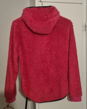 Rosa teddyjacka med huva från McKinley - Mjuk och fluffig teddyjacka i stark rosa färg från McKinley. Jackan har huva, två fickor med svarta detaljer och hel dragkedja framtill. Perfekt för kalla dagar och chill outfits. Ribbad mudd vid ärmslut och längre bakstycke för extra komfort. Det finns en fläck på ärmen