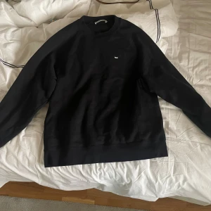 Svart sweatshirt från Wood Wood - Svart sweatshirt från Wood Wood med enkel design och liten vit logga på bröstet. Tröjan har rund hals, långa ärmar och ribbade muddar vid ärmslut och nederkant. Tillverkad i mjuk bomull som är skön att ha på sig. Har ett litet hål bakom armen, inget som stör. Original pris: 1500 kr. Perfekt julklapp till killen!