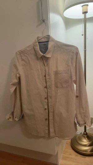Beige skjorta från WE Suiting Yourself - Snygg beige skjorta från WE Suiting Yourself i storlek 158/164. Klassisk krage, bröstficka och knappar framtill. Långärmad med möjlighet att vika upp ärmarna. Perfekt för dig som gillar enkel och stilren look.