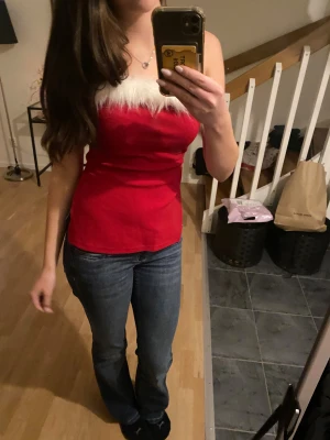 Röd jul topp - Säljer en röd jul topp med en vit fluffig överdel, perfekt för julens alla tillfällen. Toppen är axelbandslös och har en snygg slits på sidan som ger en cool detalj. Materialet känns mjukt och stretchigt, och toppen sitter tight mot kroppen💞