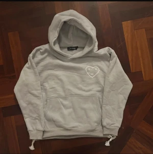 Grå hoodie med hjärtdetalj Carsicko - Säljer en ljusgrå/ljusblå hoodie från Carsicko i 100% bomull. Den har en stor huva, magficka och ett broderat hjärta med smiley och stjärnor på bröstet. Muddar vid ärmslut och nederkant samt dragsko i midjan. Perfekt för chill dagar.