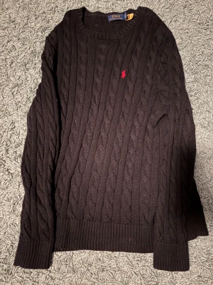 Svart stickad Ralph Lauren Tröja - Svart kabelstickad tröja från Polo Ralph Lauren med klassisk röd broderad logga på bröstet. Tröjan har har en rundad hals och passar sig som gillar denna typ av kläder. Använd ett fåtal gånger ca 5-10 gånger.