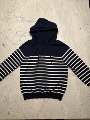 Marinblå och vit randig stickad hoodie - Snygg stickad hoodie i marinblått med vita ränder. Tröjan har ribbad nederkant och snörning vid huvan med en mjuk och skön känsla tack vare det stickade materialet. Tröjan är i storlek S och det är bara att skriva om ni har några flera frågor