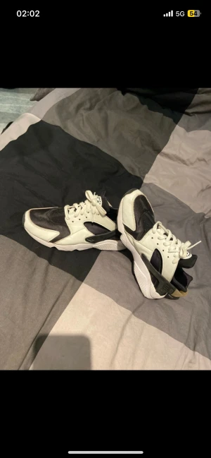 Nike Air Huarache sneakers svart/vit - Nike Air Huarache sneakers i svart och vit med detaljer i beige. Skorna har en chunky sula, snörning och en mix av syntet och skinnmaterial. Ikonisk Huarache-logga på plösen och AIR-detalj på hälen. Perfekt för dig som gillar streetwear och sportig stil.