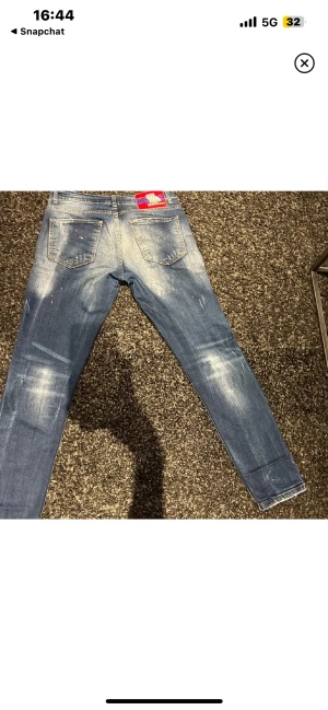Dsquared2 blå skinny jeans med slitningar - Säljer ett par blå jeans från Dsquared2 med coola slitningar, färgstänk och en röd patch bak i midjan. Jeansen har 4 fickor, låg midja och är tillverkade i stretchig denim för en tight passform. Perfekta för dig som gillar streetstyle och detaljer.