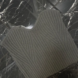 Mörkgrå ribbad stickad tubtopp - Säljer en mörkgrå stickad tubtopp med ribbad struktur och snygg bandeauform. Toppen är ärmlös och har en mjuk, varm känsla som passar perfekt till lager-på-lager. Den har en stilren look med subtila diagonala ribbmönster för extra edge.