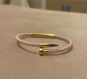 Rosa armband med gulddetaljer - Snyggt armband med flätad ljusrosa band och coola guldfärgade metalldetaljer. Designen har en unik spetsig avslutning som ger en modern vibe. Perfekt för dig som gillar stilrena accessoarer med en twist.