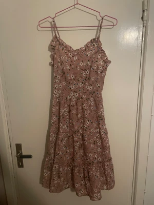 Blommig rosa sommarklänning med volang - Söt rosa klänning med vita och blå blommor, smala axelband och volangkant upptill. Klänningen har smock i ryggen för skön passform och ett luftigt, flowigt fall med volang nedtill. Perfekt för varma dagar och ger en romantisk vibe.