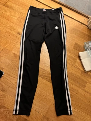 Svarta Adidas leggings med vita ränder - Svarta leggings från Adidas med klassiska vita ränder längs benen och Adidas-logga på höften. Sitter tight och har elastisk midja, perfekta för träning eller chill. Materialet är mjukt och stretchigt syntet som andas.