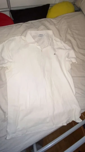 Vit piké från J.Lindeberg - Vit pikétröja från J.Lindeberg med klassisk krage och korta ärmar. Tröjan har två knappar framtill och en liten broderad logga på bröstet. Perfekt för en clean och stilren look.