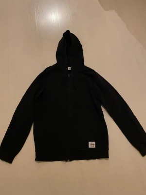 Svart hoodie från Andy by Frank Dandy - Svart hoodie med dragkedja från Andy by Frank Dandy i storlek M. Tröjan har huva med snörning, två fickor framtill och en enkel patch med logga nedtill. Materialet är mjuk bomull och passformen är normal. Perfekt för chill dagar.