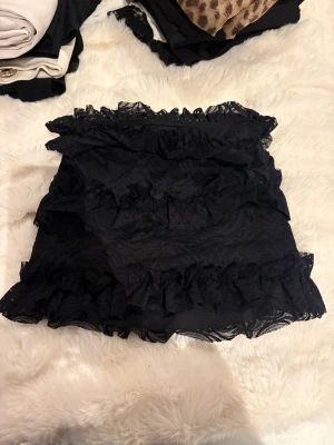 Svart volangkjol från OW - Supersöt svart kortkjol från OW med flera lager volanger och spetsdetaljer. Kjolen har en kortare tight passform och är gjord i ett syntetmaterial med transparent spets över hela ytan.