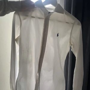 Vit skjorta från Polo Ralph Lauren - Vit långärmad skjorta från Polo Ralph Lauren i slim fit-modell. Klassisk krage, knappar framtill och den ikoniska blå broderade loggan på bröstet. Skjortan är gjord i mjuk bomull och har en stilren, tidlös look som passar till många olika outfits.