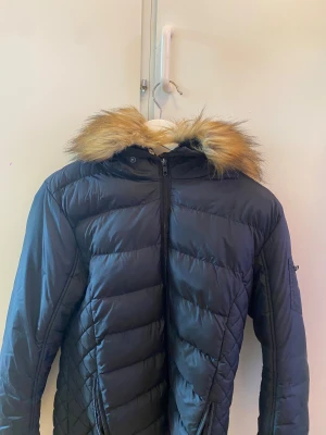 Svart pufferjacka med fuskpäls från Vinson - Svart pufferjacka från Vinson med snyggt quiltad design och huva med fluffig fuskpäls i beige/brun ton. Jackan har dragkedja framtill och två sidofickor med dragkedja. Perfekt för kalla dagar och har en modern look som passar till streetwear.