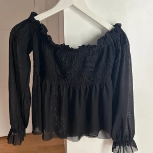 Svart blus med smock från Gina Tricot - Supersöt svart blus från Gina Tricot i storlek XS. Blusen har smock upptill, volang vid ärmslut och en luftig, transparent känsla. Offshoulder-modell med långa ärmar och rynkade detaljer som ger en romantisk vibe.