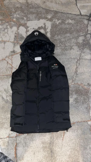 Svart dunjacka från Sail Racing - Svart dunjacka från Sail Racing med huva och dragkedja framtill. Jackan har en stilren design med logotyp på bröstet och är perfekt för kalla dagar. Fylld med dun och har en längre passform som ger extra värme. Snygg och funktionell för vintern.