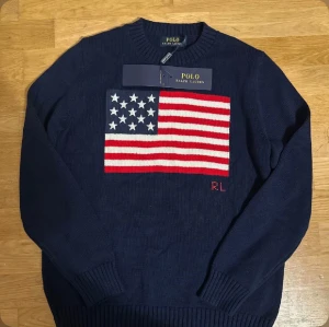 Mörkblå stickad tröja Polo Ralph Lauren - Snygg mörkblå stickad tröja från Polo Ralph Lauren med amerikansk flagga i vitt och rött på bröstet samt RL-broderi. Rund hals och ribbade muddar vid ärmslut och nederkant. Perfekt för dig som gillar klassisk och cool stil.