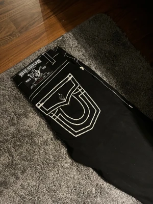 Svarta True Religion jeans med kontrastsömmar - Svarta jeans från True Religion med vita kontrastsömmar och ikoniska fickdetaljer bak. Klassisk femficksmodell med raka ben och silverfärgade knappar. Snyggt broderad logga på bakfickan och patch i midjan. Skriv pm om frågor och mer bilder. Priset kan diskuteras. Tveka inte på att skriva 