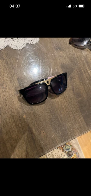 Louis Vuitton svarta solglasögon guld - Snygga solglasögon från Louis Vuitton med svart båge och guldiga detaljer på sidorna och vid näsryggen. Rektangulär form och mörka glas. Tillverkade i Italien, perfekt för dig som vill sticka ut med en lyxig accessoar.