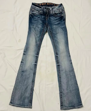 Rock Revival bootcut jeans blå - Snygga blå bootcut jeans från Rock Revival med slitningar och detaljerade bakfickor med broderi och knappar. Jeansen har låg midja och klassisk femficksdesign. Perfekt för dig som gillar en cool och avslappnad stil.