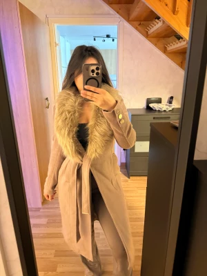 Beige kappa med fuskpäls och bälte - Snygg beige kappa med bred krage i fuskpäls och knytbälte i midjan. Jackan har långa ärmar med  guldfärgade knappar vid ärmslutet. Bra skick. 