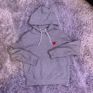 Grå hoodie Comme des Garçons Play M - Grå hoodie från Comme des Garçons Play i storlek M. Tröjan har huva med vita snören och ett broderat rött hjärta med ögon på bröstet. Klassisk kanguruficka framtill och mjukt bomullsmaterial. Perfekt för chill dagar och streetwear vibes. Obs den säljes billigt för att det finns småa fläckar på framsidan. 