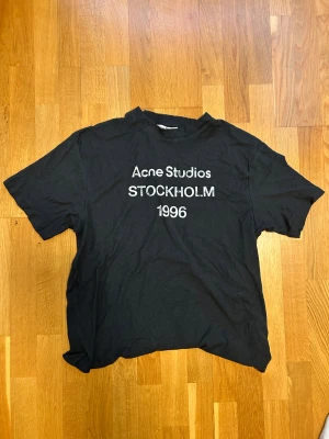 Svart Acne Studios t-shirt - Svart t-shirt från Acne Studios med vit text 'Stockholm 1996' på bröstet. Klassisk rund hals och korta ärmar. Materialet är mjuk bomull och passformen är normal. Perfekt för dig som gillar stilrena och ikoniska plagg med streetkänsla. Storleken är Xs men passar S. Den här två små hål vid kragen men syns knappt