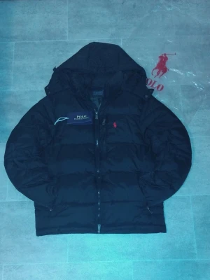 Svart pufferjacka Polo Ralph Lauren - Svart pufferjacka från Polo Ralph Lauren med röd broderad logga på bröstet. Jackan har huva, tvåvägsdragkedja och quiltad design. Perfekt för kalla dagar och har en clean, sportig look. Har kvitto.