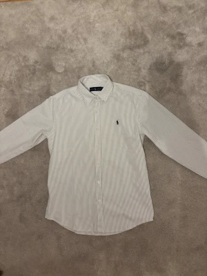 Randig skjorta från Ralph Lauren - Säljer en riktigt snygg Ralph Lauren Skjorta i Stolek L. Skjortan är i utmärkt skick. Säljer den eftersom jag tyckte att den var för stor för mig. Färgen är vit med gråa ränder