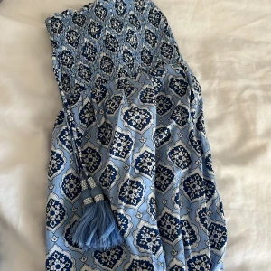 Blå långkjol  från Gina Tricot - Säljer en blå långklänning från Gina Tricot med ett snyggt blommigt mönster i mörkblått och vitt. Klänningen har smock upptill och dekorativa tofsar i midjan. Perfekt för sommarens alla tillfällen, superfräsch och bohemisk vibe.