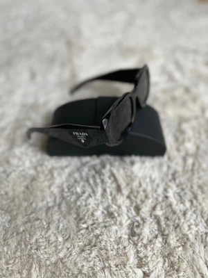 Svarta solglasögon från Prada - Snygga svarta solglasögon från Prada med rektangulär form och breda skalmar. Glasögonen har en stilren design med diskret logga på sidan och kommer med originalfodral och original låda och certifikat. Perfekta för dig som vill ha en lyxig och modern look.