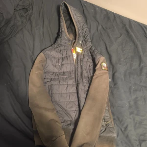 Parajumpers svart pufferjacka S - Svart pufferjacka från Parajumpers med huva och coolt patch på ärmen. Jackan har quiltad framsida, dragkedja, fleeceärmar och flera fickor. Insidan har säkerhetsficka och orange detaljer. Perfekt för dig som gillar streetwear och vill ha något varmt och snyggt. 