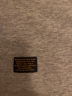 Grå sweatshirt Morris R. Club L - Snygg grå sweatshirt från Morris R. Club i storlek L. Tröjan har ett broderat emblem med brittisk flagga och gulddetaljer på bröstet. Klassisk rund hals och långa ärmar med ribbade muddar. Perfekt för dig som gillar stilren och sportig design.