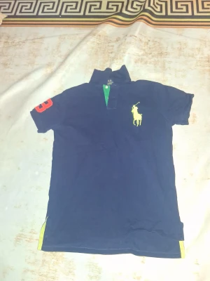 Mörkblå Polo Ralph Lauren t-shirt - Snygg mörkblå t-shirt från Polo Ralph Lauren med klassisk polokrage och gul logga på bröstet. Röd siffra 3 på ärmen och gröna samt gula detaljer vid kragen. Custom fit-modell i mjuk bomull, perfekt för dig som gillar sportig och stilren look.