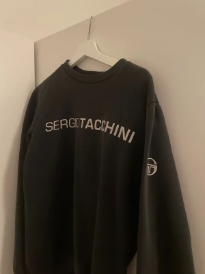 Svart sweatshirt från Sergio Tacchini - Svart sweatshirt från Sergio Tacchini i storlek S. Tröjan har ett stort tryck med loggan över bröstet och en mindre logga på ärmen. Rund hals och lång ärm, tillverkad i mjuk bomullsmix för skön komfort. Perfekt för en chill och sportig look.