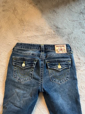 True Religion Becca Bootcut Jeans - Snygga blå jeans från True Religion, modell Becca Mid Rise Bootcut. Klassisk femficksdesign med gul kontrastsöm och ikoniska ficklock bak med knappdetaljer. Jeansen har bootcut-ben och midwaist. Materialet är slitstarkt denim med coola detaljer. Lite slitna längst ner💗💗